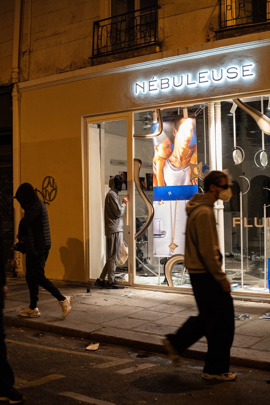 Vitrine de bijouterie fracturée la nuit à Paris, un individu encapuchonné à l'intérieur, des passants sur le trottoir