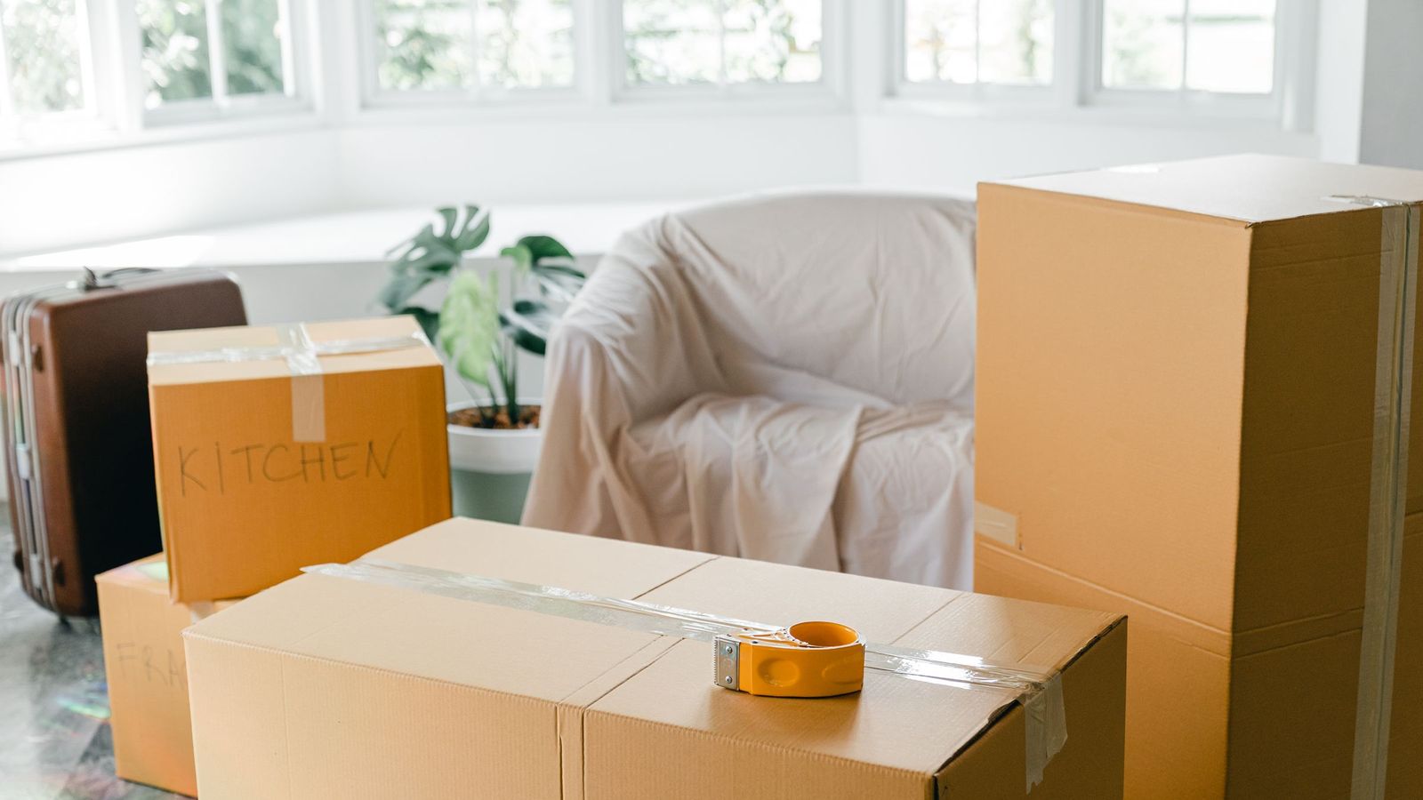 Cartons de déménagement et fauteuil protégé par un drap dans un séjour lumineux bordé de grandes fenêtres